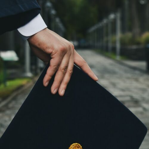 A close-up shot of a hand holding a graduation cap on an outdoor path, symbolizing achievement.