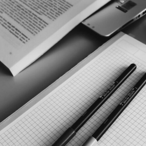 Black and white closeup of a study desk with an open book, grid notebook, and pens for a minimalist study scene.